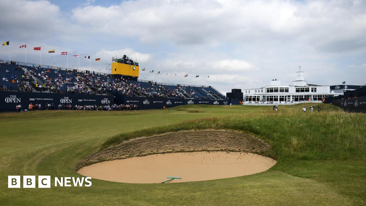 a golf course, with a sandy bunker in the foreground. On the left there is a large stand, approximately half full of fans. Above that, there is a small TV gantry for cameras. To the right of the stand, a large white building is pictured. On the course, there are several golf players and caddies in the distance.