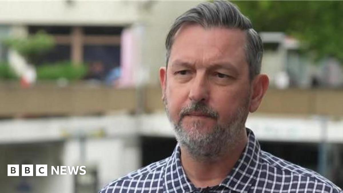 Chris has grey hair and a grey beard. He is looking just off camera and is wearing a chequered blue and white shirt.