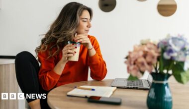 Woman with long, light brown hair wearing an orange long-sleeved top and black leggings sits at a table at home drinking a smoothie and looking at a laptop
