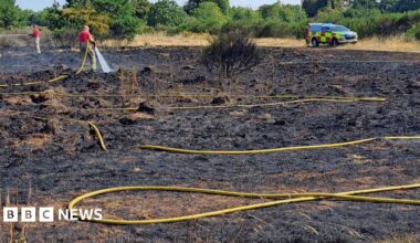 Two firefighters in red shirts and yellow trousers are holding long yellow hoses and spraying water onto scorched ground after a grass fire. In the background is a fire service vehicle parked on unburnt grass with trees and a blue sky behind.