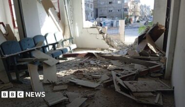 A badly damaged wall of an office, debris is scattered inside it and dust covers a bank of witting room chairs