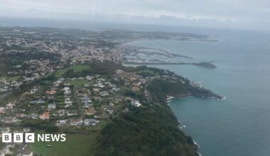 Arial view of Guernsey. The sea is a dark blue and the sky is grey and full of clouds.