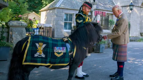 PA Media King Charles III - who is wearing tweed jacket, matching waistcoat, white shirt and blue tie; kilt, black shocks and black shoes - meets Shetland pony Cpl Cruachan IV (mascot of the Royal Regiment of Scotland) during an inspection of the Balaklava Company, 5th Battalion, The Royal Regiment of Scotland, at the gates of Balmoral, as he takes up summer residence at the castle. 