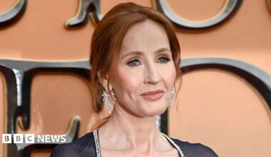 Head shot of Harry Potter author JK Rowling, She has red hair, tied back in a ponytail and is wearing long diamond earrings. She is wearing a black dress, with a diamond trim. Rowling is standing in front of a pink backdrop advertising "Fantastic Beasts: The Secret of Dumbledore". She is looking off to the left of the camera.