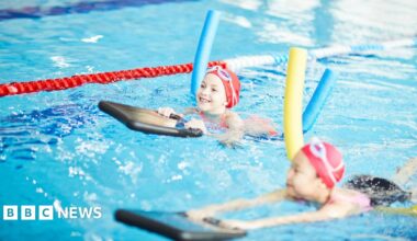 Two young girls are swimming in a pool. They are both wearing red swimming caps and goggles. Each of them is holding a black float and are using a pool noodle, one blue and one yellow.