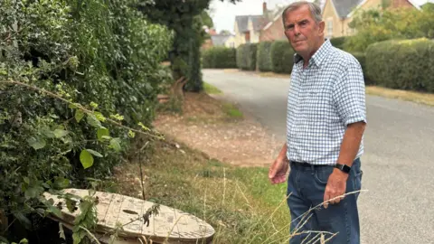BBC A man, wearing is a blue and white check shirt and blue jeans, is standing on the roadside looking at a wooden reel of cables next to the hedge.   