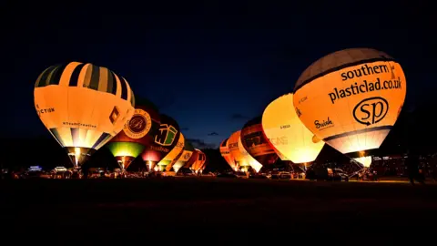 Getty Images Two lines of hot air balloons tethered to the ground. They are all lit up from their burners. The image is taken at night time. 