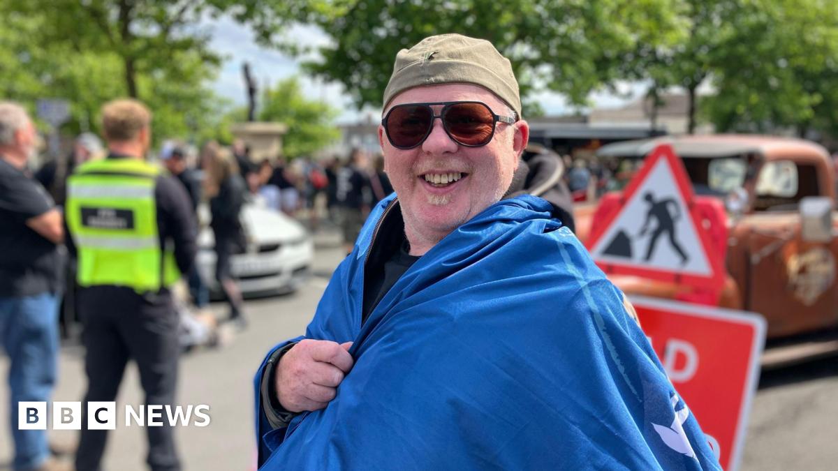 Andy Edwards, a man wearing sunglases, is draped in a blue flag. He is smiling for the camera.