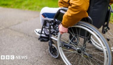 A stock image of a person sitting in a wheelchair, wheeling it along a tarmac path. The image shows the person from the elbow down, they are wearing an orange raincoat and grey tracksuit bottoms.