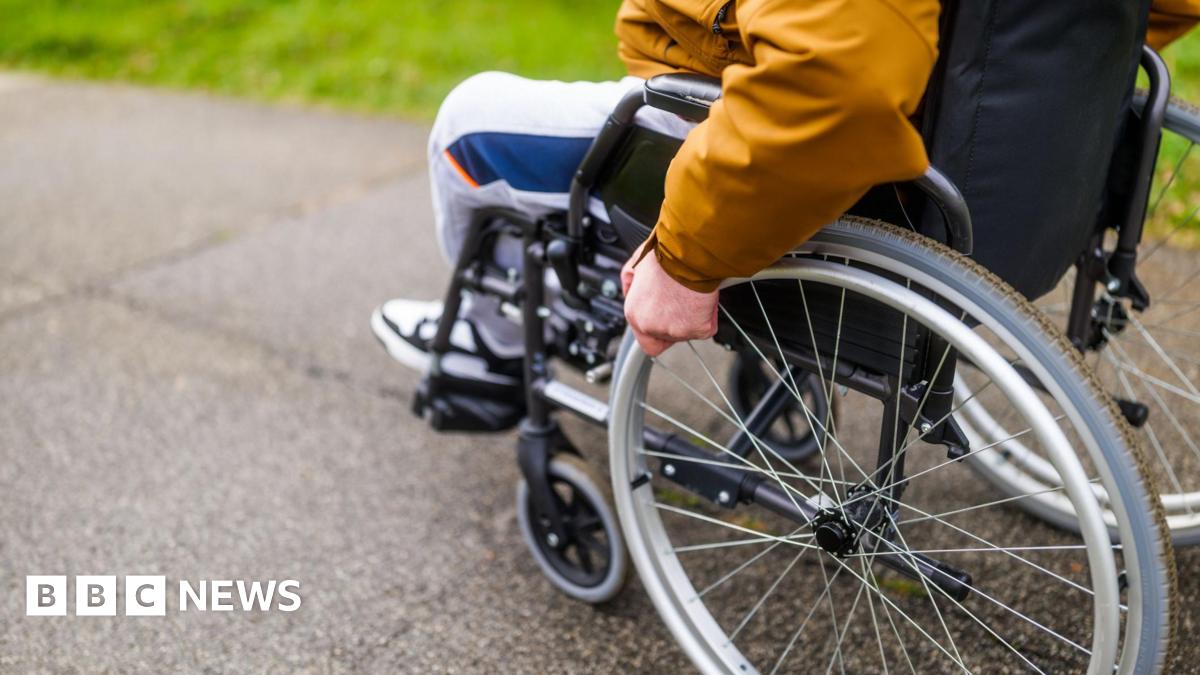 A stock image of a person sitting in a wheelchair, wheeling it along a tarmac path. The image shows the person from the elbow down, they are wearing an orange raincoat and grey tracksuit bottoms.
