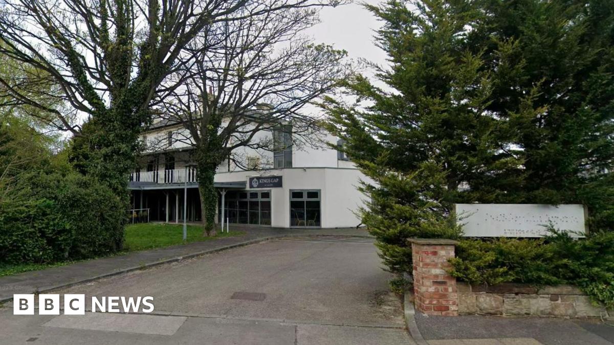 A white two-storey hotel building behind a low brick wall entrance with grass and trees on either side. There is a white sign at the entrance where the former hotel name has been removed.