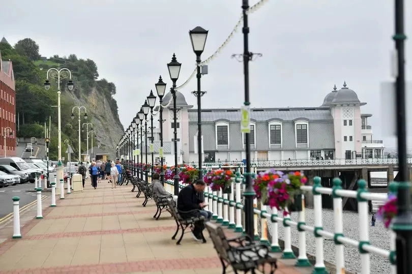 People walking on a pavement on the right, with the Penarth Pier and beach visible on the left