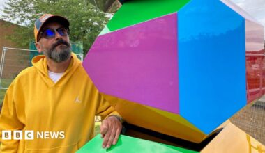 A man in a yellow hooded top and baseball cap looking at a multi-coloured block.