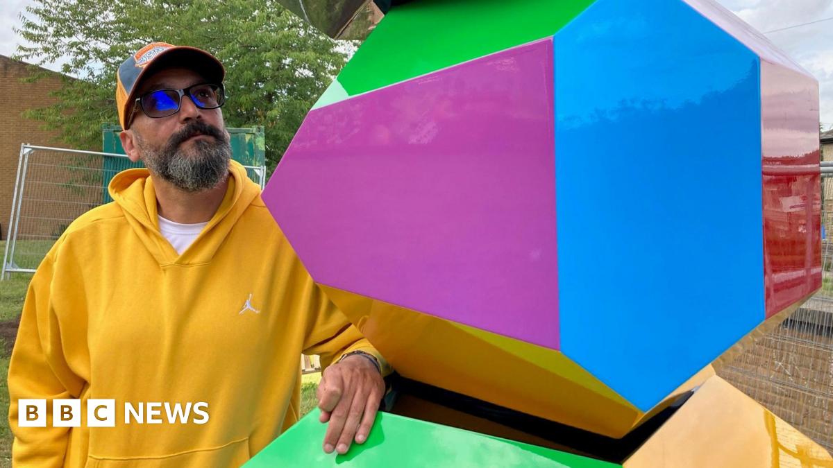 A man in a yellow hooded top and baseball cap looking at a multi-coloured block.