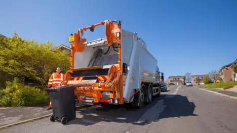 Dorset Council A white and orange bin collection lorry stopped at the side of a residential street. A man in orange overalls stands at the back with a black wheelie bin. It's a sunny day and the sky is blue.