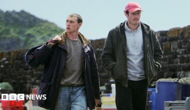 George MacKay and Callum Turner in character are walking towards the camera. Mullion's granite sea wall is behind them and the walkway is littered with barrels of rope, nets, piled up fish trays and mooring buoys. MacKay has short blond brown hair. He has a sports bag slung over his right shoulder and is wearing pale blue jeans, a sweatshirt and a heavy coat. He is looking right with a serious expression. Turner is wearing a faded red cap over his short dark brown hair and is wearing black sweatpants, a pale grey hoodie and a dark grey zip up hoodie over the top. He has a faint goatee and is looking down with his lips pursed.