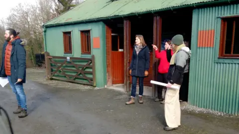 Robjn Cantus Fiona Bruce with members of the Fake or Fortune crew coming out of a green-painted metal barn. She is standing in a doorway on the right, wearing blue jeans, a blue jacket, gloves and her light brown hair is should-length. She is looking to the left, in the direction being pointed to by a woman in a red fleece. A member of the crew on the left wearing a hat, jacket and cream trousers is looking towards her. Another member of the crew with a dark beard and hair in a bun, wearing a blue jacket and jeans is on the far left. 