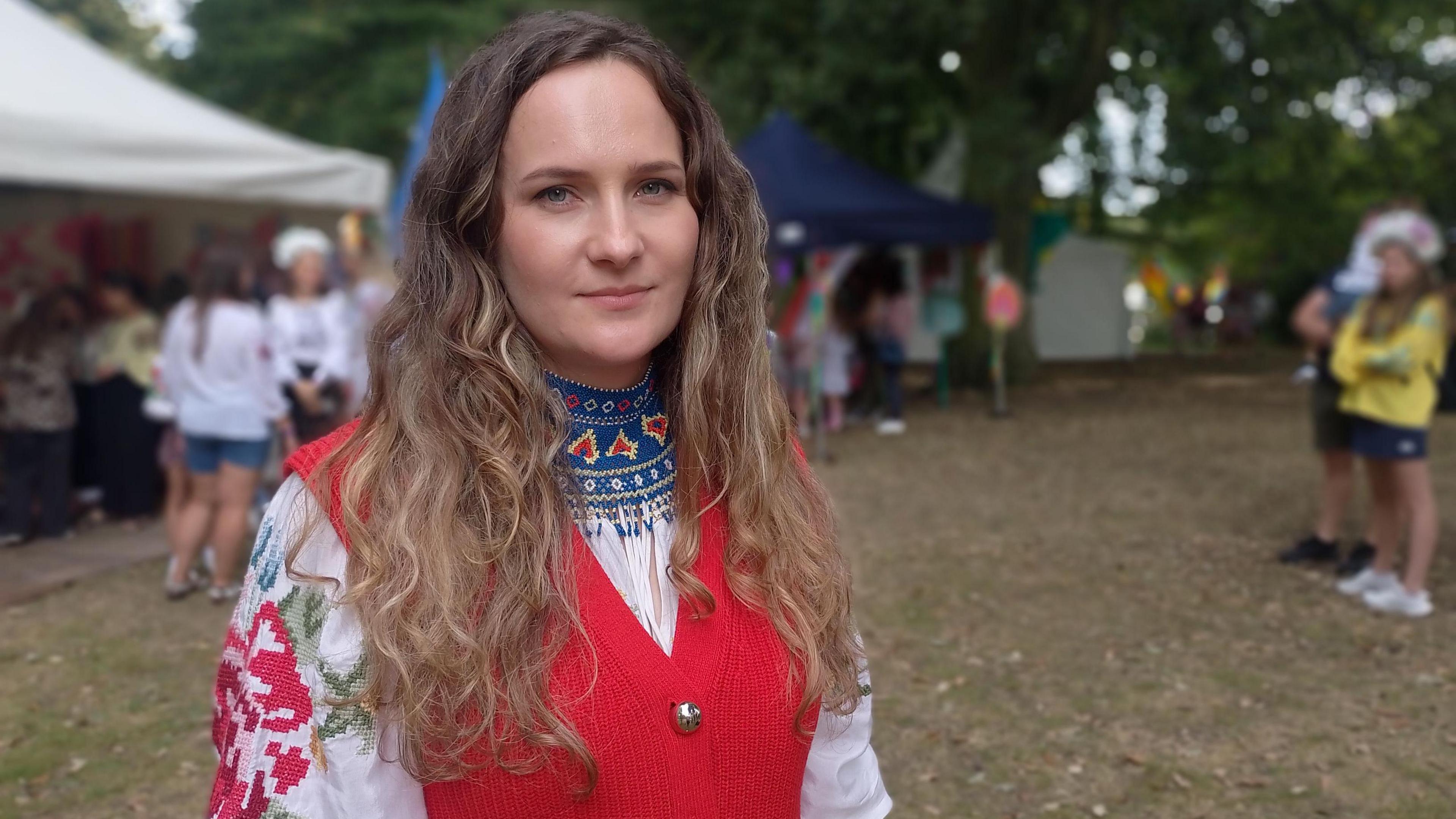 A woman with light brown curly long hair wears traditional clothes. They are an embroidered white top with a red wasitcoat. 