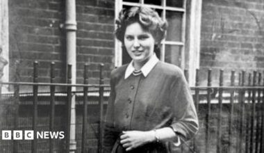 A black and white photo of a young woman with short dark curly hair. She wears a cardigan with white collar sleeves and stands in front of a brick building with a black fence.