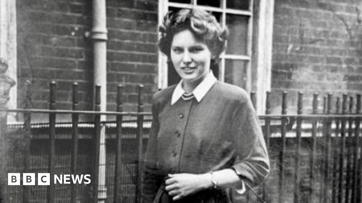 A black and white photo of a young woman with short dark curly hair. She wears a cardigan with white collar sleeves and stands in front of a brick building with a black fence.