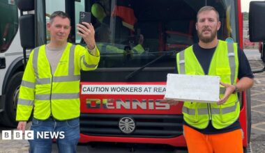 Two workers for Barnsley's waste collection service, stood in front of a bin lorry, wearing high visibility jackets. One holds aloft a mobile phone. The other holds the technology used to track mobile signal coverage as the lorries traverse the area - which looks like a white box.