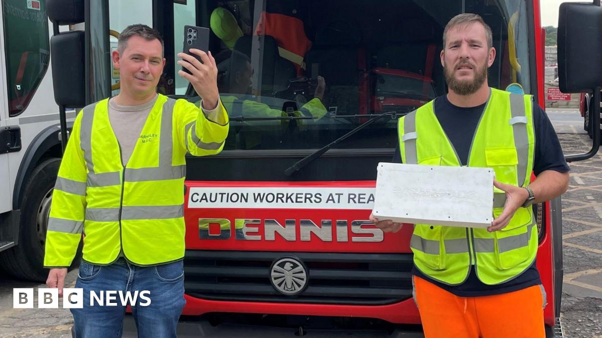 Two workers for Barnsley's waste collection service, stood in front of a bin lorry, wearing high visibility jackets. One holds aloft a mobile phone. The other holds the technology used to track mobile signal coverage as the lorries traverse the area - which looks like a white box.