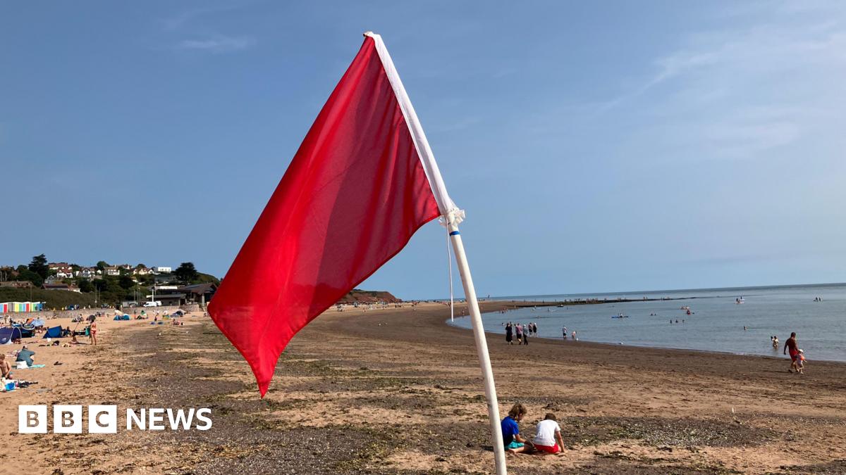 A red flag is blowing in the wind on a beach. Beachgoers are in the distance. Swimmers are in the water. The sky is blue.