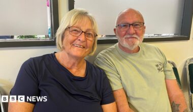 A head and shoulder shot of a woman and a man - Bea and Frank Carter - sitting down smiling at the camera. Bea has blonde, bobbed hair and is wearing glasses and a navy t-shirt. Frank is bald and has grey facial hair and is wearing glasses and a green t-shirt. He has a tattoo of a star on his left arm.