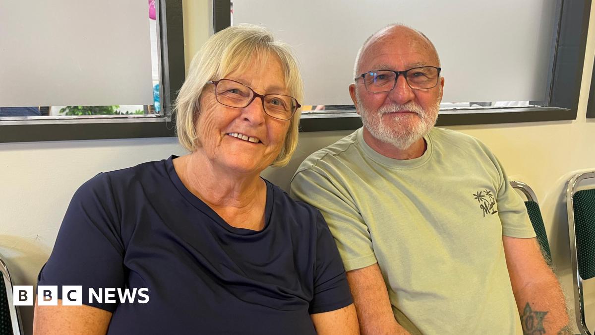 A head and shoulder shot of a woman and a man - Bea and Frank Carter - sitting down smiling at the camera. Bea has blonde, bobbed hair and is wearing glasses and a navy t-shirt. Frank is bald and has grey facial hair and is wearing glasses and a green t-shirt. He has a tattoo of a star on his left arm.