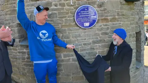 James Atkin wearing a blue tracksuit and cap, with his right hand in the air as he smiles down to Ian Dench who is wearing all black and a blue beanie. They are holding a cloth together which they have just taken from the blue plaque on the stone wall behind them. The blue plaque is in recognition of their music achievements. 