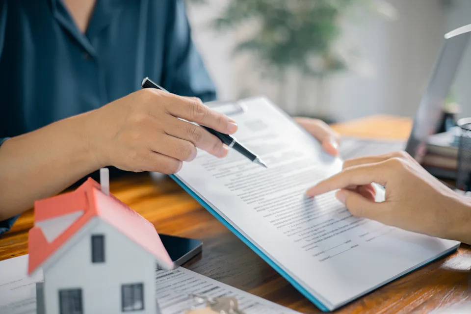 Two people discuss documents at a desk; one holds a pen over paperwork while the other points. A small house model is on the table