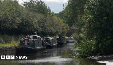 At least three narrowboats are in the background. There are trees on both sides of the canal.