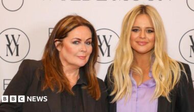 Kate Robbins, with brown hair, is stood next to Emily Atack, with blonde hair, at a press event. There is a white wall behind them with a brand logo that looks like a W and M joined together in a circle.
