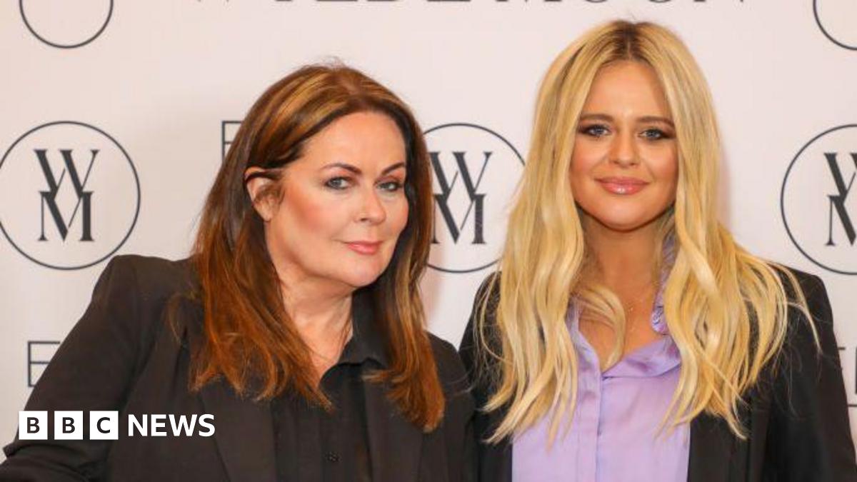 Kate Robbins, with brown hair, is stood next to Emily Atack, with blonde hair, at a press event. There is a white wall behind them with a brand logo that looks like a W and M joined together in a circle.