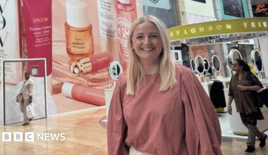 A blonde woman wearing a pink shirt smiling inside of a shopping centre