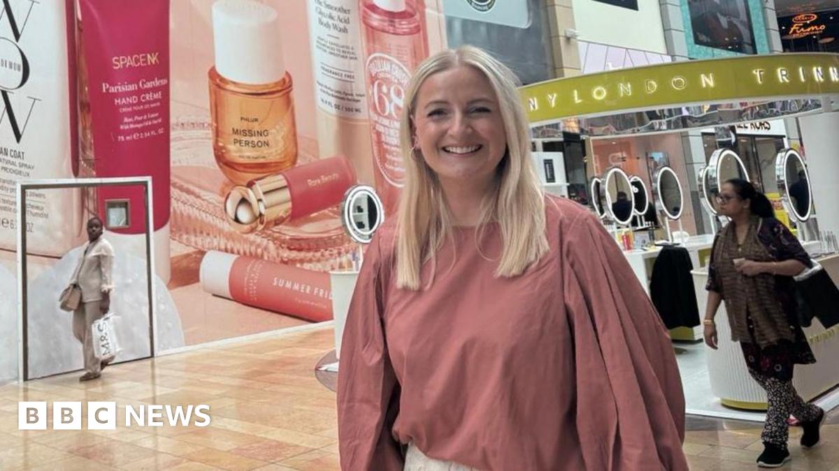A blonde woman wearing a pink shirt smiling inside of a shopping centre