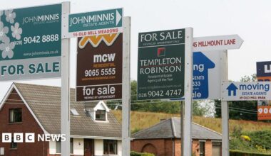 A row of 'For Sale' signs in front of two houses.