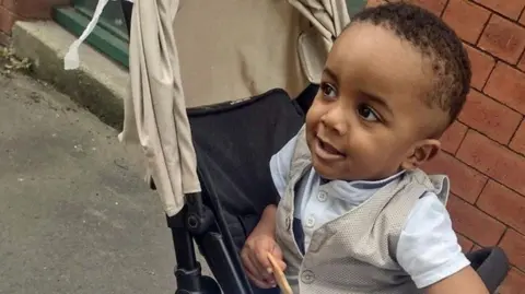 Family pic Family photo of Exodus Eyob, a small boy looking to the side of the camera while sitting in a black pushchair with a cream hood. He has short dark hair and is wearing a white t-shirt with buttons at the neck and a cream waistcoat, and is holding a biscuit. He is pictured outside with a brick wall behind him.