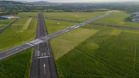 Gloucestershire Airport Ltd Gloucestershire Airport's two tarmac runways, which cross in an X shape. Around the runways are dark green fields.