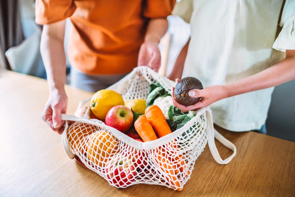 Close-up shot of a mother and daughter unpacking a reusable mesh bag filled with fresh fruits and vegetables in the kitchen. The focus is on the hands holding the bag and an avocado, emphasising the importance of healthy eating and sustainable practices. This scene highlights the themes of family bonding, healthy lifestyle and eco-friendly living.