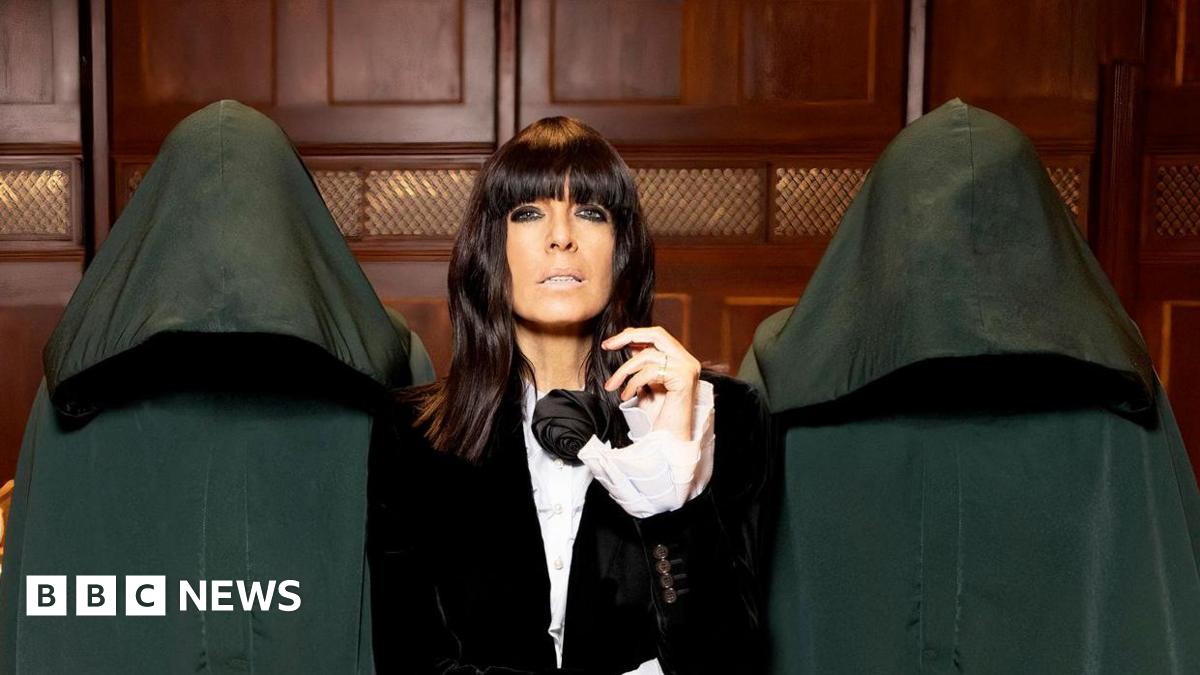 Claudia Winkleman wears a dark suit and white shirt. She has brown hair that goes just past her shoulders and wears heavy eyeliners. She is flanked by two anonymous people in hooded cloaks. They are in a wood-panelled room