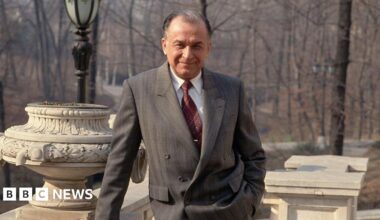 Iliescu poses and looks straight down the camera with a slight smile. He is leaning on a stone balcony and wearing a grey suit.