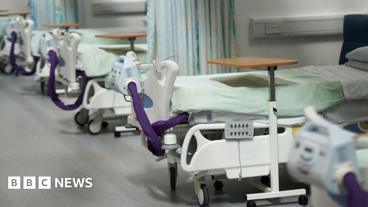 A stock image of a row of inpatient beds at a hospital. They are empty with a swivel table over the bed and medical equipment attached to the plastic frames. Each bed is separated with a drawn-back curtain.
