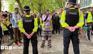 Police with their backs to the camera holding a line of police in front of counter-protestors. There is a large number of counter-protestors begin one lady how is shouting into a megaphone.