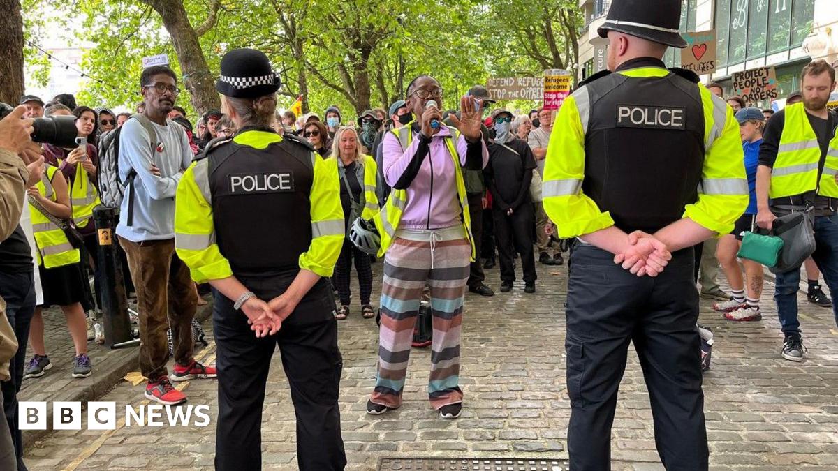 Police with their backs to the camera holding a line of police in front of counter-protestors. There is a large number of counter-protestors begin one lady how is shouting into a megaphone.