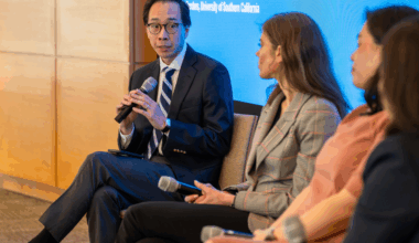Two women and one man in professional attire sit on a panel on stage and speak using microphones.