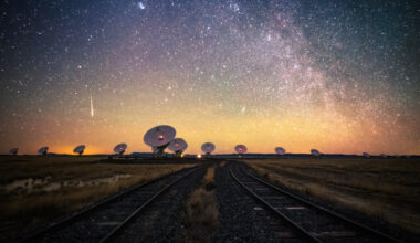 a dozen large white radio telescopes sit beneath a starry sky with train tracks in the foreground