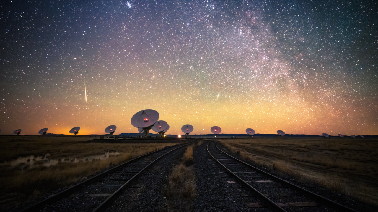 a dozen large white radio telescopes sit beneath a starry sky with train tracks in the foreground