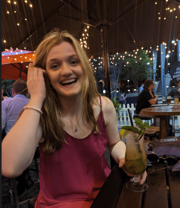 A smiling young woman with long blonde hair is sitting at a table, holding a cocktail decorated with mint leaves and a slice of lime, wearing a pink top. The background features string lights and other patrons in a lively outdoor setting.