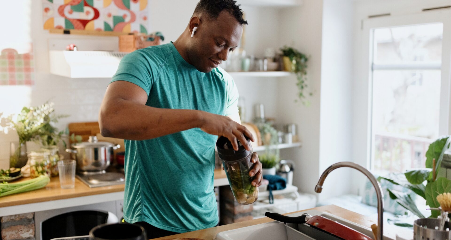 man in a turquoise tshirt in a kitchen by the sink holding a blender cup screwing the lid on.